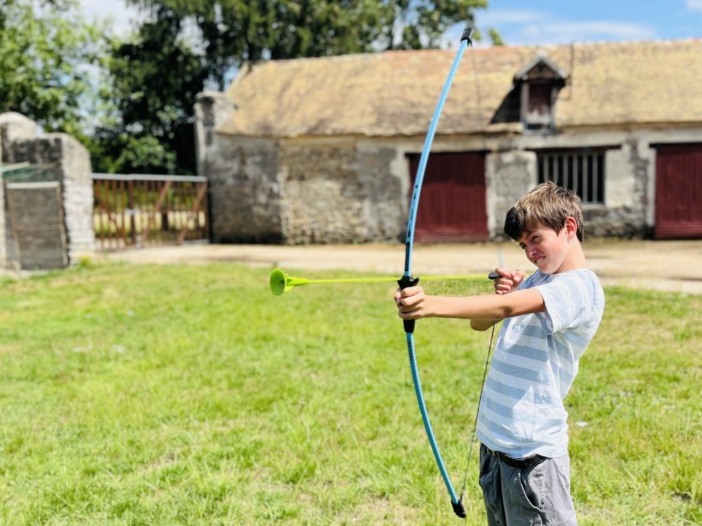 Un enfant vise avec un arc lors d'une activité de plein air, avec les bâtiments en pierre de la ferme de loisirs en arrière-plan