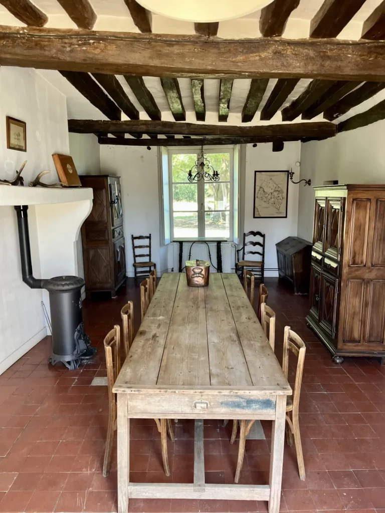 Décor de cinéma authentique à Bruyères-le-Châtel (Essonne) : salle à manger de la Ferme de La Forêt avec poutres apparentes, sol en tomettes, poêle à bois et mobilier rustique. Idéal pour tournages de films d'époque et shootings photo proche Paris